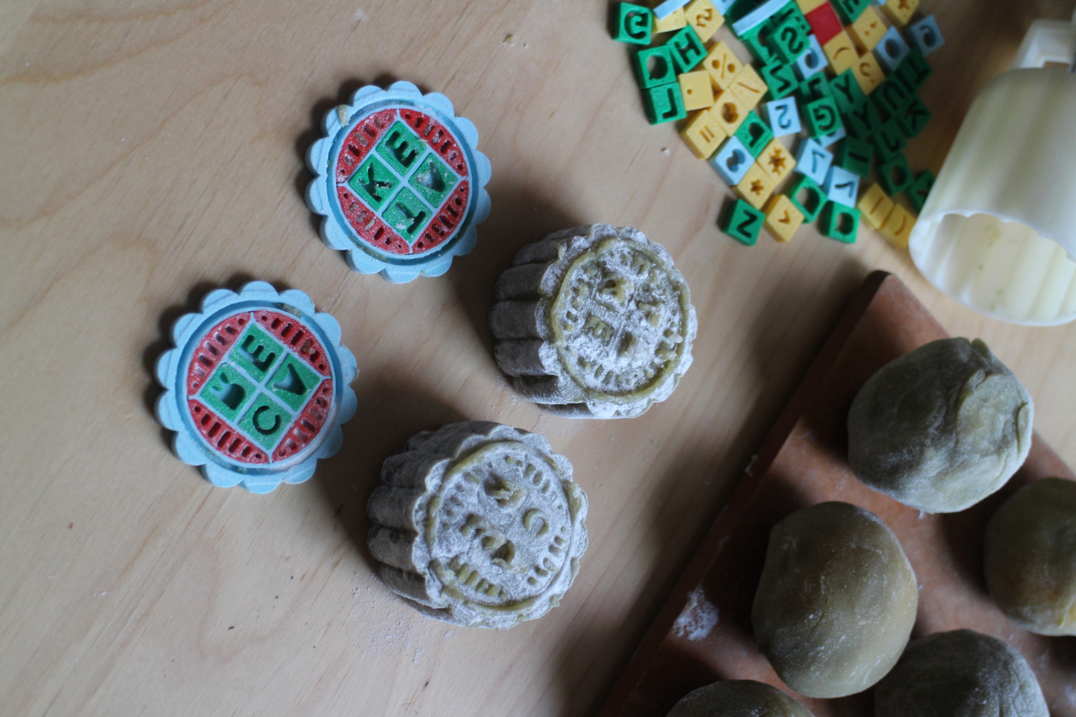 Two mooncakes with the words 'TAKE CARE'  imprinted on their surface by customizable mooncake stamps. The mooncake stamps are customizable with 3D printed letters, numbers, symbols. Balls of mooncake dough in the background.