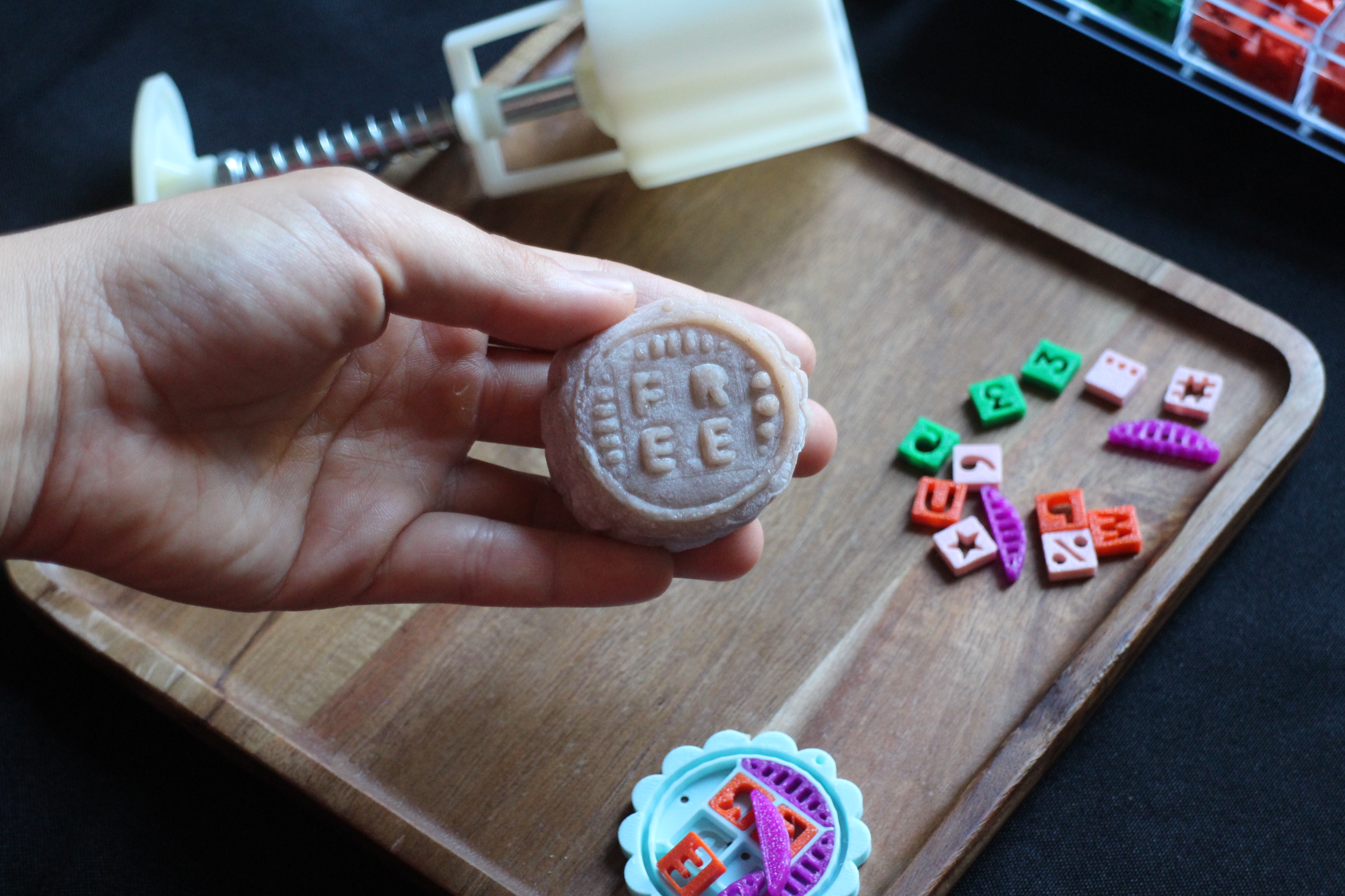 A light-skinned hand holding a pink mooncake with the letters 'FREE' stamped on it. In the background, there is a mooncake stamp, and colorful 3D printed letters and designs.