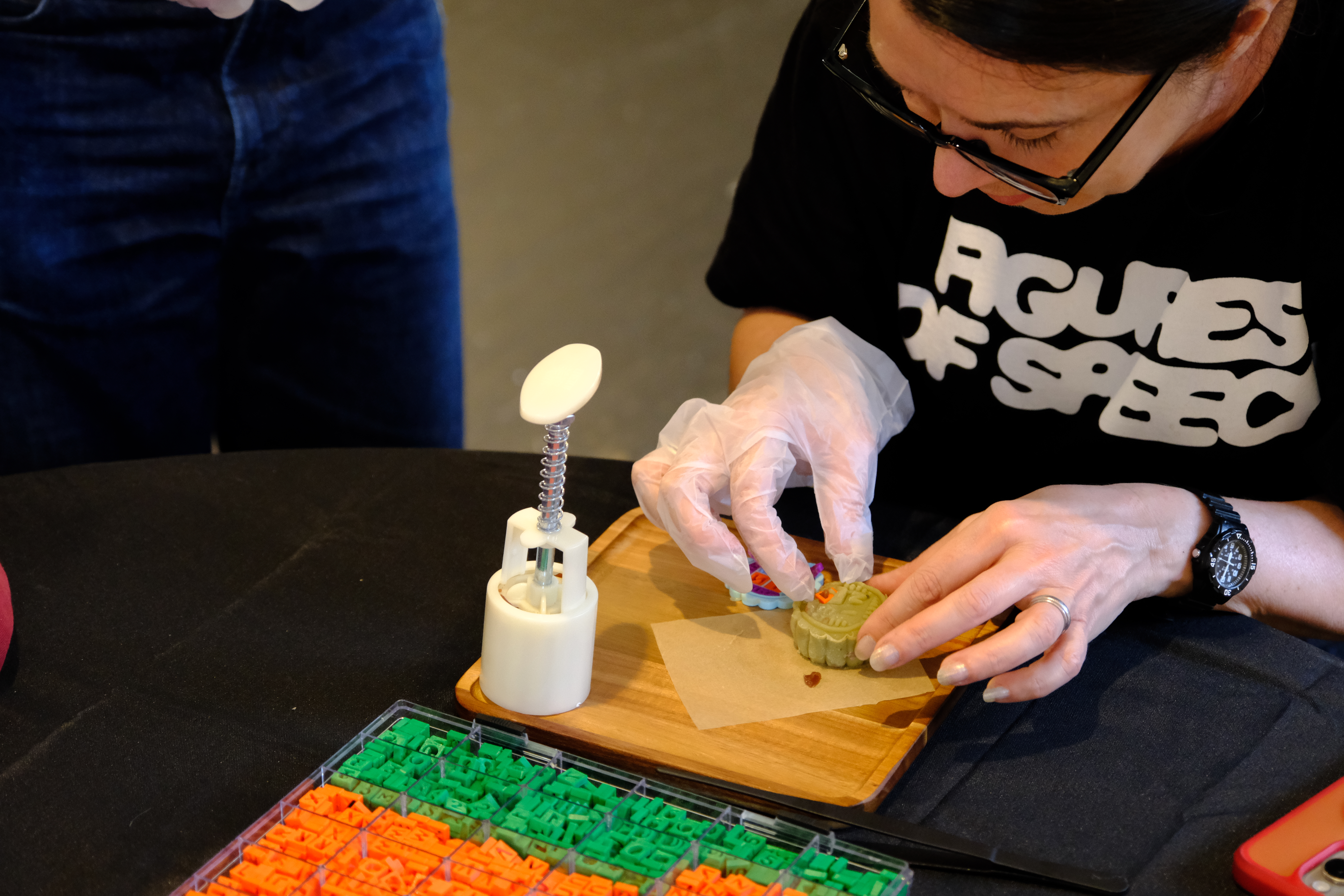 A white woman with black hair sitting down, creating her PROTEST MOONCAKE with 3D printed designs and a green matcha mooncake. There is a mooncake stamp and colorful 3D printed letters and designs in front of her.